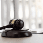 Brown wooden gavel resting on a circular base with a blurred background of light-colored vertical blinds.