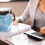 A blue piggy bank with a graduation cap sits on a stack of papers next to a calculator, as a person writes nearby.