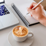 Cup of latte with leaf design beside a laptop and a hand holding a pen above a blank notepad.