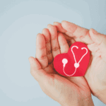 Red paper heart with a white stethoscope design held between two hands against a light blue background.
