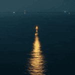 Lone buoy with a bright light in the ocean at night, reflecting golden light on the water surface. Distant lights visible on the horizon.