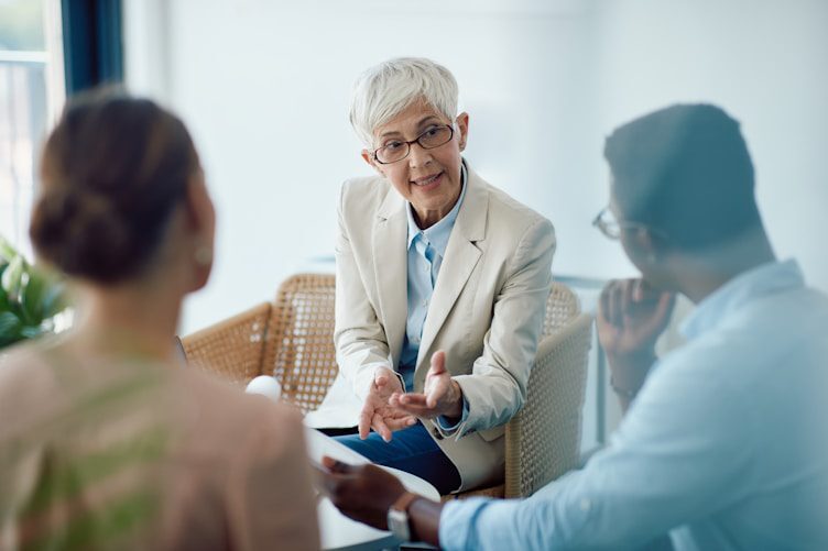 A woman with short gray hair in a beige blazer gestures while speaking to two seated individuals in a bright, modern room.
