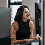 Smiling woman with glasses and arm tattoos sitting at a desk, facing a computer screen, in a modern office setting.