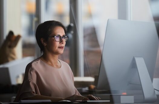 Woman with short hair and glasses sitting at a desk, focused on a computer screen, with large windows in the background.