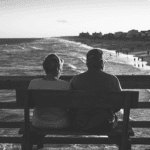 Elderly couple sitting on a bench, facing the ocean. Waves and beach visible in the background, creating a serene sunset atmosphere.