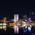 Rochester skyline at night, featuring illuminated buildings and a bridge over a reflective river.