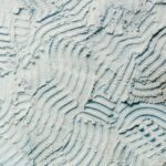 Light blue surface with textured, wavy patterns created by tire or tool marks. Dusty appearance with fine particles.