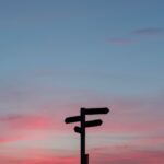 Silhouette of a signpost with multiple arrows against a colorful sunset sky with pink and blue hues.