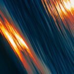 Sunset reflections on water, with streaks of orange and gold against deep blue waves, creating a dynamic, abstract effect.