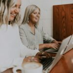 Two women smile while working on laptops at a wooden table. One woman has long, light hair and wears a white top; the other has straight, light hair and is dressed in a striped blouse. A coffee cup is placed on the table.