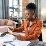 Young Black woman in a brown polka dot shirt sitting at a desk, reviewing documents with a calculator and smartphone nearby.