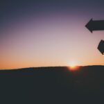 Silhouette of a signpost with multiple arrows against a sunset, with the sun just above the horizon and a gradient sky.
