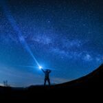A person stands on a hill at night, pointing a flashlight towards the starry sky, with the Milky Way visible.