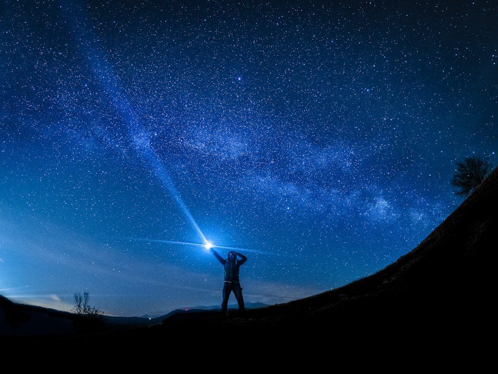 A person stands on a hill at night, pointing a flashlight towards the starry sky, with the Milky Way visible.