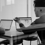 Two people sit at a table with laptops, discussing work. The scene is captured through a glass partition, creating a blurred effect.