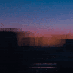 Blurred city skyline at sunset with vibrant pink, orange, and blue hues; silhouettes of buildings and cranes visible.