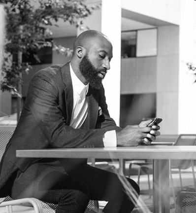 Man with a beard sitting at a table outdoors, looking at his phone. Modern building and greenery in the background.