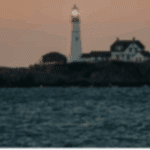 Lighthouse on rocky shore at sunset, with water in the foreground and a soft glow from the light atop the tower.