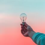Hand holding a clear light bulb against a gradient pink and blue sky background.
