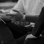 Two men are seated on a bench, one wearing a white shirt and a watch, gesturing while holding a tablet. The other is in a black suit.