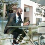A man with a beard sits at a metal table in an outdoor area, using his phone. He wears a dark blazer, white shirt, and sneakers.