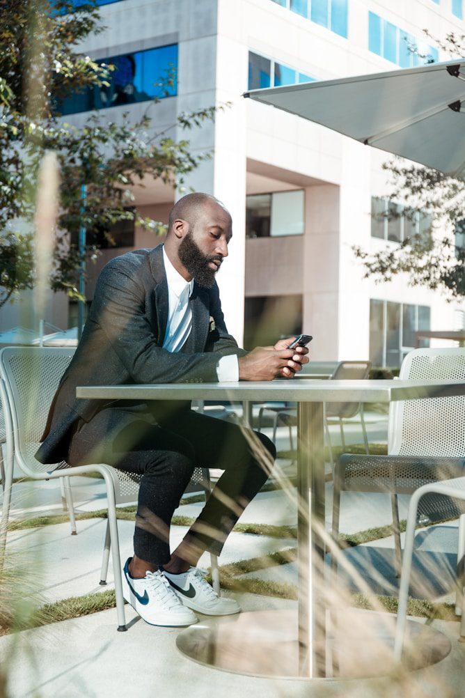 A man with a beard sits at a metal table in an outdoor area, using his phone. He wears a dark blazer, white shirt, and sneakers.