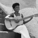 Smiling young woman with short curly hair playing an acoustic guitar while sitting on stone steps.