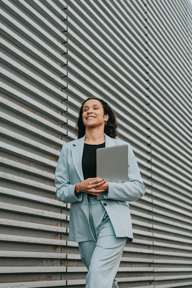 Smiling woman in a light blue suit holding a laptop, walking beside a striped metal wall.