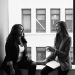 Two women sit by a large window, smiling and engaging in conversation. One wears a black blazer, and the other has a gray blazer with a laptop.
