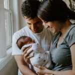 A smiling couple holds a swaddled newborn near a window, with a world map in the background.