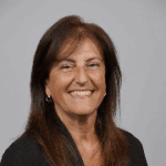 Smiling woman with long brown hair, wearing a black jacket, against a gray background.