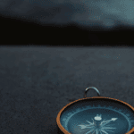 A close-up of a vintage compass with a dark blue dial and orange rim, resting on a textured gray surface, with a blurred background.