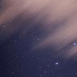 Starry night sky with scattered stars against a dark blue background, partially obscured by wispy clouds.