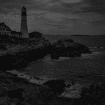 A rocky coastline features a tall lighthouse beside a dark house, under a cloudy sky, with waves crashing against the rocks.