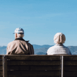 Two elderly individuals sit on a wooden bench, facing away, with mountains and a clear blue sky in the background.