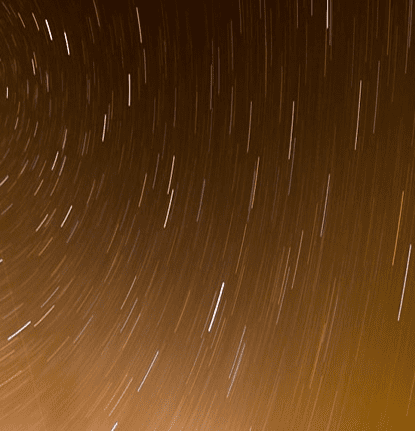 Long exposure photo showing a starry night sky with streaks of light creating circular patterns against a dark orange background.