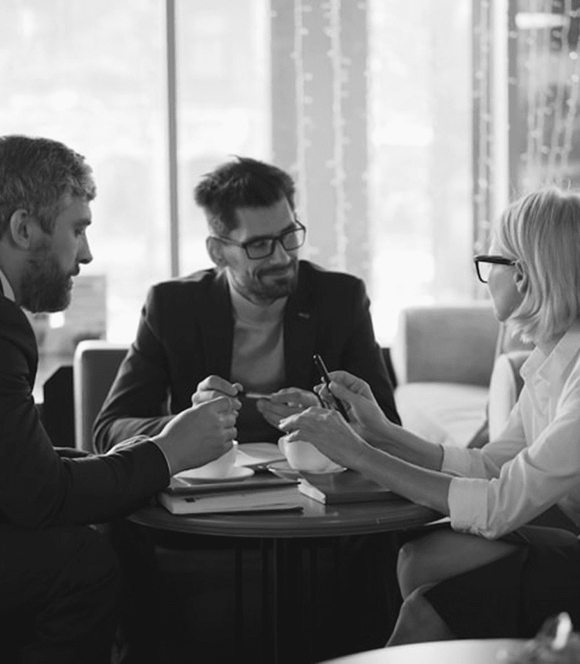 Three professionals engaged in a discussion at a table, with a coffee cup and notepad. The setting is bright, with soft lighting.