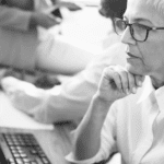 A thoughtful older woman with short gray hair and glasses sits at a desk, resting her chin on her hand, focused on a computer.