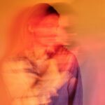 A woman with a thoughtful expression is partially blurred against an orange background, creating a sense of motion.