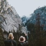 Two hikers with backpacks stand facing a rocky mountain landscape, one pointing towards a distant waterfall amid the trees.