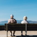 Two elderly individuals sit on a bench, facing away, overlooking a scenic view of mountains and water under a clear blue sky.
