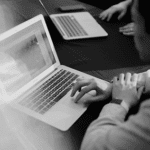 A person with short dark hair is using a laptop, focusing on the screen. Another laptop is visible on the table.