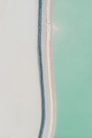 Aerial view of a shoreline with a sandy beach in light beige, bordered by dark gray rocks, and turquoise water.