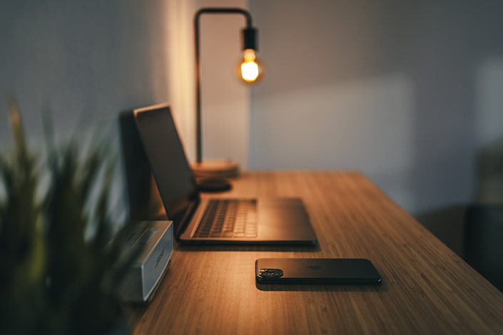 A wooden desk with a laptop, a smartphone, and a lamp casting warm light, with a plant in the background.