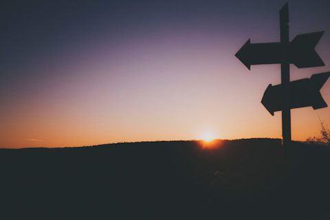Silhouette of a signpost with multiple arrows against a sunset, with the sun just above the horizon and a gradient sky.