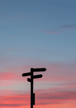Silhouette of a directional signpost against a colorful sunset sky with pink and blue hues.