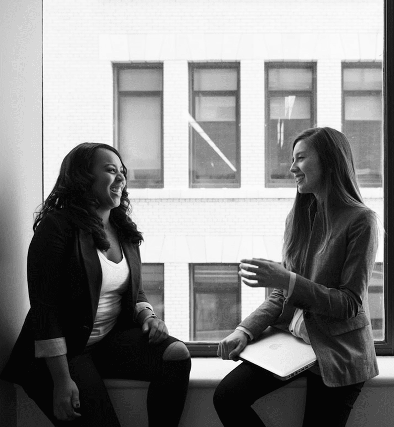 Two women sit by a large window, smiling and engaging in conversation. One wears a black blazer, and the other has a gray blazer with a laptop.
