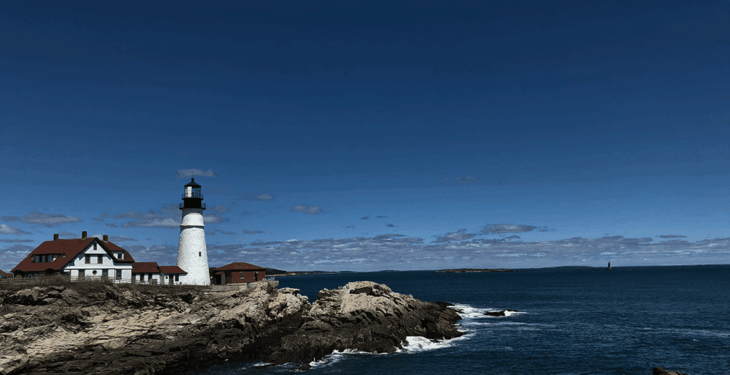 Lighthouse with a white tower and red-roofed building on rocky coast, under a blue sky with scattered clouds, ocean waves visible.