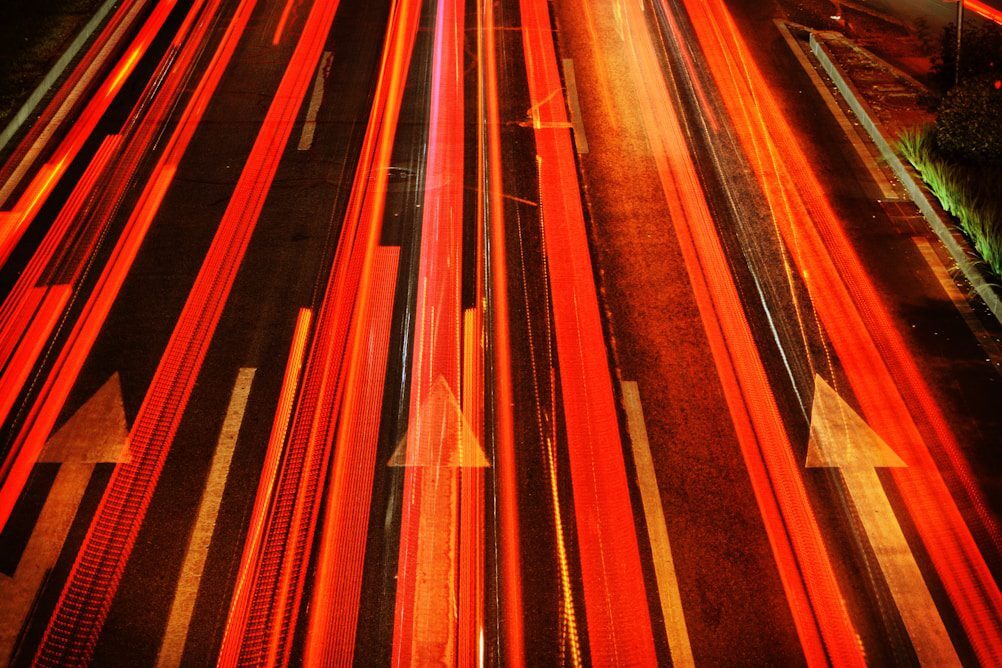 Bright red and orange light streaks from moving vehicles on a dark road with white arrow markings indicating direction.
