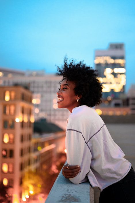 Smiling woman with curly hair leans on a rooftop railing, overlooking a city skyline at dusk with buildings illuminated.